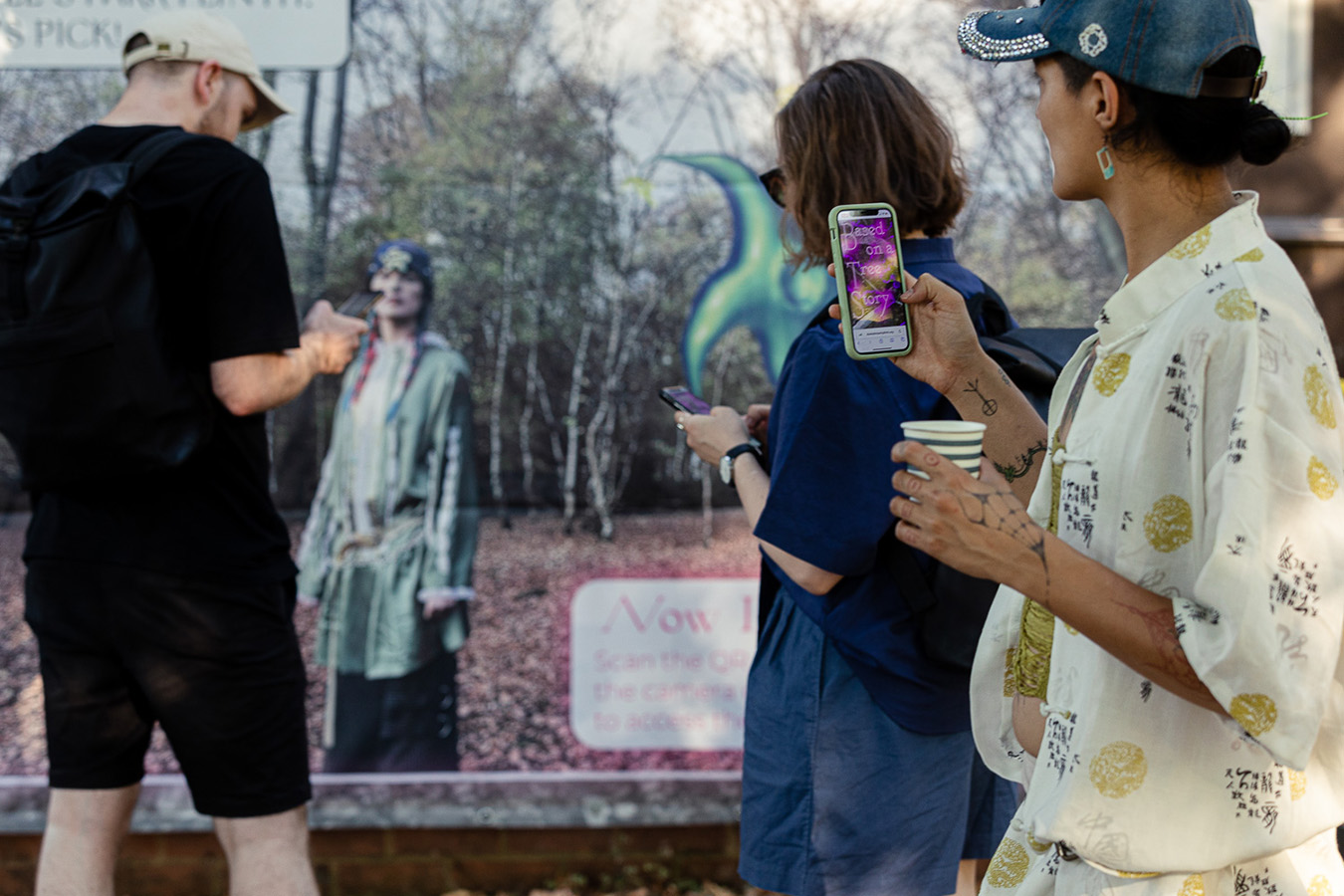 3 people in front of a sign, using their phones, based on a tree story can be read on the closest persons phone