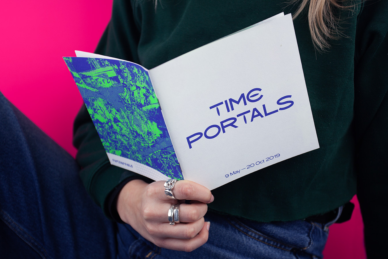 alt: A girl sat in front of a neon pink background, holding a booklet which reads Time Portals on the front cover and has a neon blue and green image on the back cover.