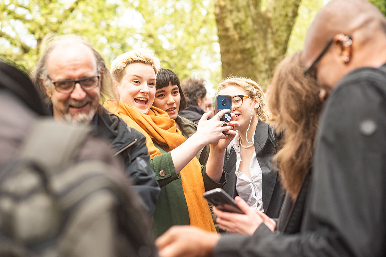 3 people in the background laughing while looking at a phone, while 4 people in the foreground out of focus laugh